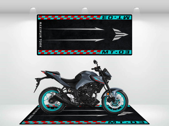 In a showroom, a black and gray motorcycle with turquoise accents stands on a Motorcycle Pit Mat for Yamaha MT-03, featuring MT-03 and Maximum Torque text along with arrows and checkered patterns to enhance the bike’s presentation.