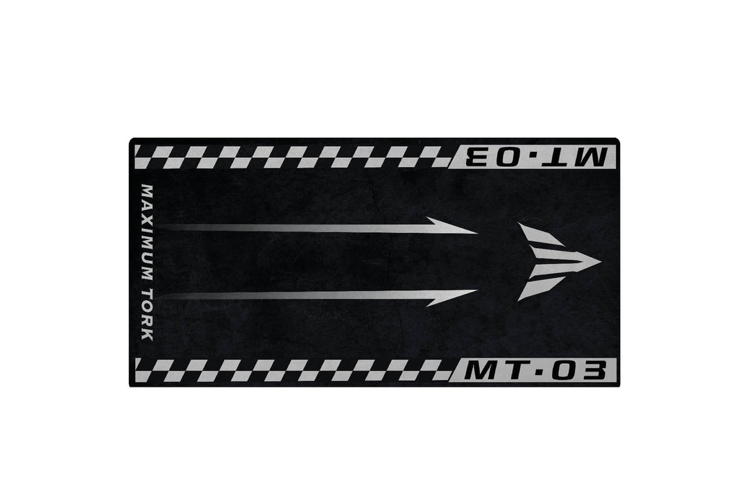 The Motorcycle Pit Mat for Yamaha MT-03 is a black and gray rectangle featuring MAXIMUM TORK and MT.03 on the edges, two upward arrows, and a stylized triangle logo. The textured background gives it a rugged look.