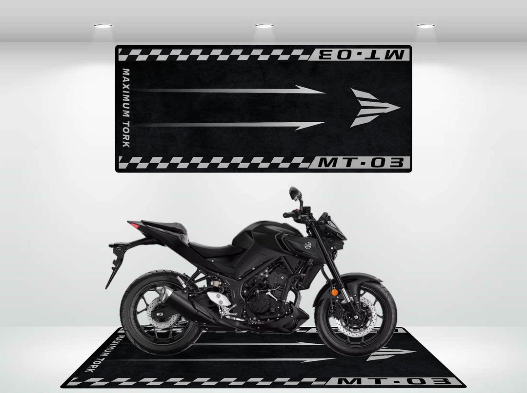 A sleek black motorcycle is showcased on a Motorcycle Pit Mat for Yamaha MT-03 with a matching wall graphic, both adorned with arrows, checkerboard patterns, and the text MT-03 and MAXIMUM TORQ, all highlighted by bright spotlights.