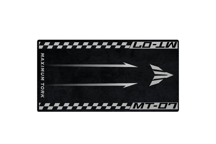 The Motorcycle Pit Mat for Yamaha MT-07 is a rectangular black mat featuring three silver arrows pointing right. MAXIMUM TORQUE is on the left edge, with MT-07 at the top and bottom. It has a tire tread design along the border.