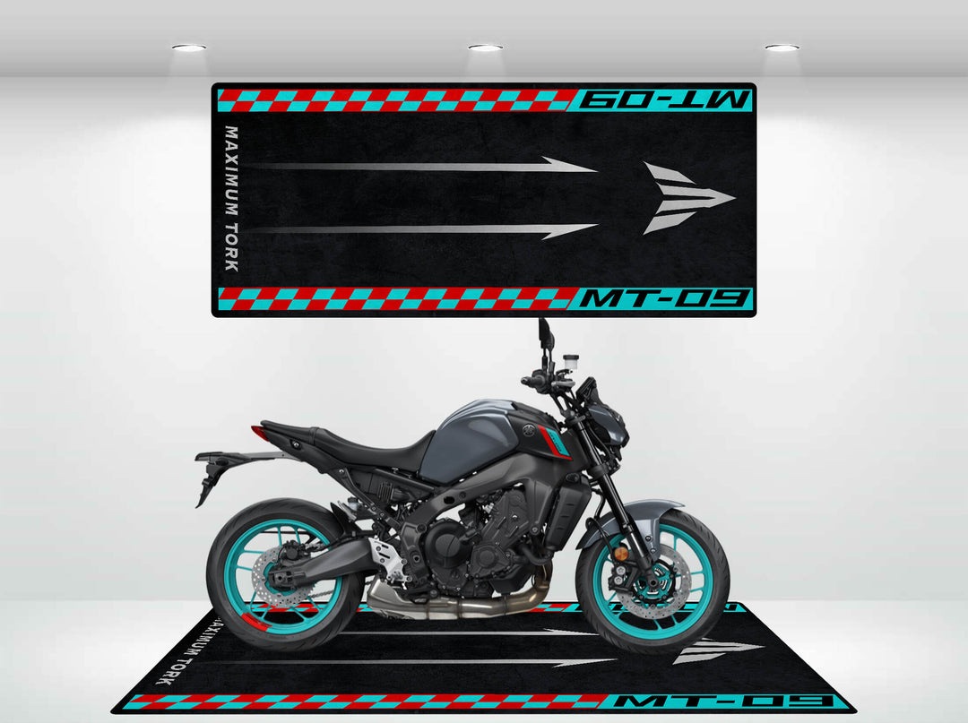 A black motorcycle with teal rims sits on a Motorcycle Pit Mat for Yamaha MT-09, featuring MT-09 and Maximum Torque. An identical mat hangs behind on a plain white wall under overhead lights.