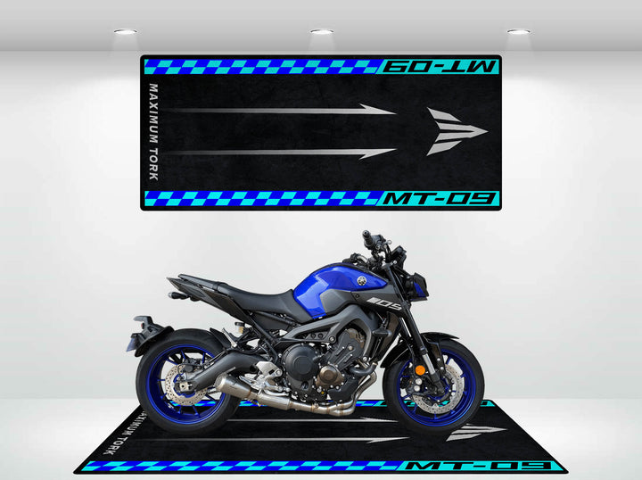 In a showroom, a blue motorcycle rests on the Motorcycle Pit Mat for Yamaha MT-09, surrounded by artwork featuring MT-09 and Maximum Torque, accented with sleek black and blue directional arrows.