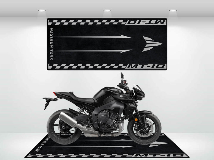 In a white showroom, a black motorcycle is displayed on a checkered Motorcycle Pit Mat designed for the Yamaha MT-10. The mat features MT-10 and MAXIMUM TORQUE with arrows, all illuminated by bright overhead lights.