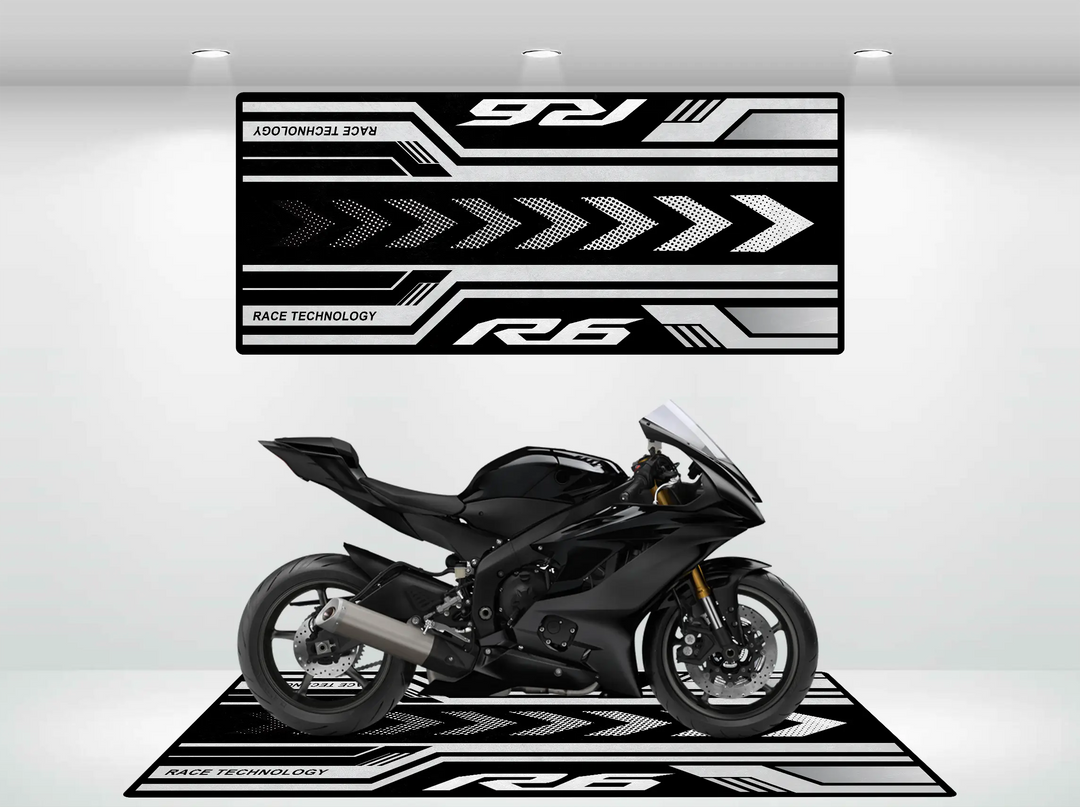 A sleek black Yamaha R6 sport motorcycle is showcased in a showroom featuring a Motorcycle Pit Mat, with black and white Race Technology graphics, bold arrows, and racing motifs on the walls. The lighting accentuates the bikes design.