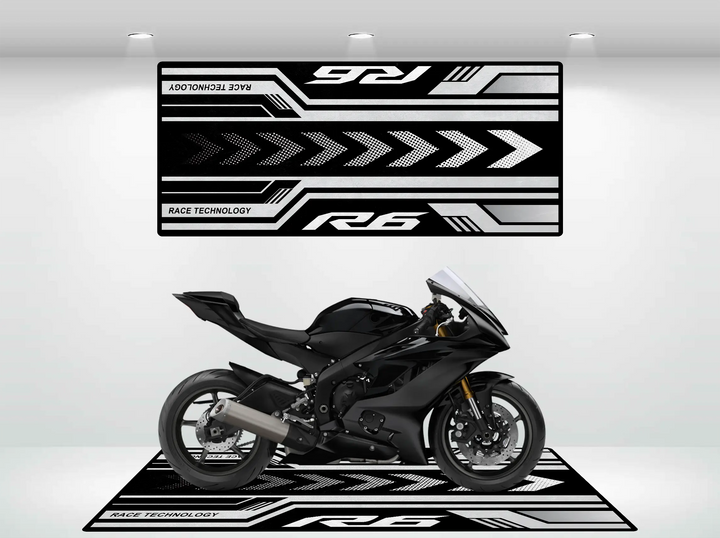 A sleek black Yamaha R6 sport motorcycle is showcased in a showroom featuring a Motorcycle Pit Mat, with black and white Race Technology graphics, bold arrows, and racing motifs on the walls. The lighting accentuates the bikes design.