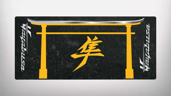 The black rectangular motorcycle mat for Suzuki Hayabusa features a yellow traditional gate design with a stylized symbol center. It has silver metallic text on both sides, creating an elegant and sleek finish, characteristic of the Motorcycle Pit Mat brand.